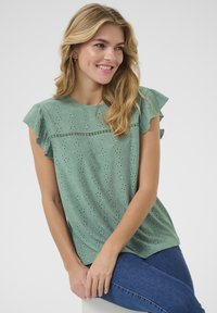 Top in cotone verde con design a occhiali, maniche arricciate, scollatura rotonda e cuciture decorative sul petto. Indossato con jeans in denim.