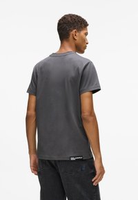 Mężczyzna odwrócony plecami, ubrany w gładką, ciemnoszaro t-shirt z małą metką "Karl Lagerfeld Jeans" oraz czarne dżinsy.