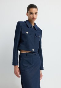 Námorně modrá zkrácená bunda se stříbrnými knoflíky, čtyřmi předními kapsami a klasickým límcem, doplněná o odpovídající sukni s vysokým pasem.