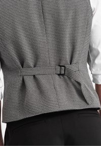 Bakvy av en person som bär en grå texturerad väst med justerbart band och spänne över en vit skjorta och svarta byxor.