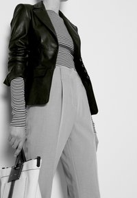 Černý kožený blejzr navrstvený přes černobílou pruhovanou košili s dlouhým rukávem, doplněný šedými skládacími kalhotami a bílou taškou přes rameno.