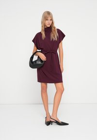 Μπορντό φόρεμα με κοντά μανίκια και ζώνη στη μέση, σε συνδυασμό με μία μαύρη τσάντα και μαύρες μυτερές γόβες, μπροστά σε ουδέτερο φόντο.