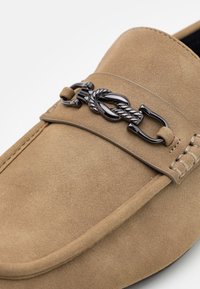 Beige suede loafers with a curved shape, featuring a black metallic chain accent and detailed stitching along the edges.