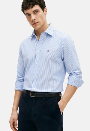 Młody mężczyzna w jasnoniebiesko-białej koszuli w paski z podwiniętymi rękawami i ciemnych spodniach, stojący z jedną ręką w kieszeni.