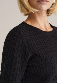Čierny textúrovaný sveter s geometrickým vzorom, okrúhlym výstrihom a rebrovanými manžetami, ktorý zdôrazňuje mäkkú a ľahkú pleteninu.