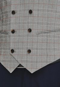 Szary w kratkę dwurzędowy kamizelka z ciemnymi guzikami, noszona na granatowych spodniach, z dwiema poziomymi kieszeniami oraz szpiczastym dołem.