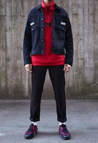 Czarna dżinsowa kurtka z kieszeniami z przodu, czerwona bluza z kapturem, spodenki w prążki oraz czarne i burgundowe sneakersy. Swobodny, warstwowy wygląd.