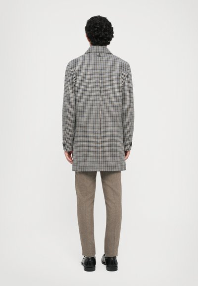 Man med krulligt mörkt hår som bär en mellanhängande houndstooth-kappa, beige byxor och svarta skor, som står vänd mot en enfärgad vit bakgrund.