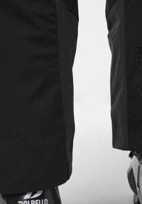 Pantalones de esquí negros con una tela texturizada, que presentan paneles contrastantes y detalles de marca. Vista cercana de las secciones laterales e inferiores.