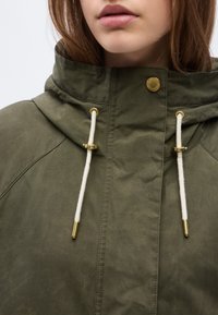 Γυναίκα που φοράει χακί μπουφάν με κουκούλα, λευκά κορδόνια και χρυσές μεταλλικές λεπτομέρειες, κοντινό πλάνο στον γιακά και τα χείλη.