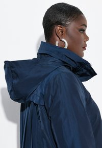 Giacca impermeabile blu navy con colletto alto, cappuccio e dettagli plissettati sul retro. Realizzata in materiale liscio e lucido con cuciture visibili.
