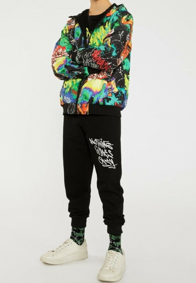 Personne debout, les bras croisés, portant une veste à imprimé graffiti coloré, un pantalon noir avec du texte graffiti blanc, des chaussettes à motifs et des baskets blanches.