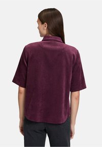 Kobieta z długimi brązowymi włosami, ubrana w krótką, ciemnofioletową, aksamitną bluzkę i czarne spodnie, stojąca plecami na białym tle.