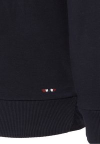 Tessuto a maglia blu navy scuro con orlo a coste e piccolo logo ricamato in rosso e bianco vicino al bordo inferiore.