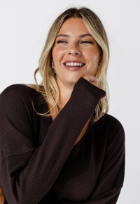 Femme blonde souriante avec des cheveux ondulés portant un pull texturé sombre, la main près de son visage, sur fond blanc uni.