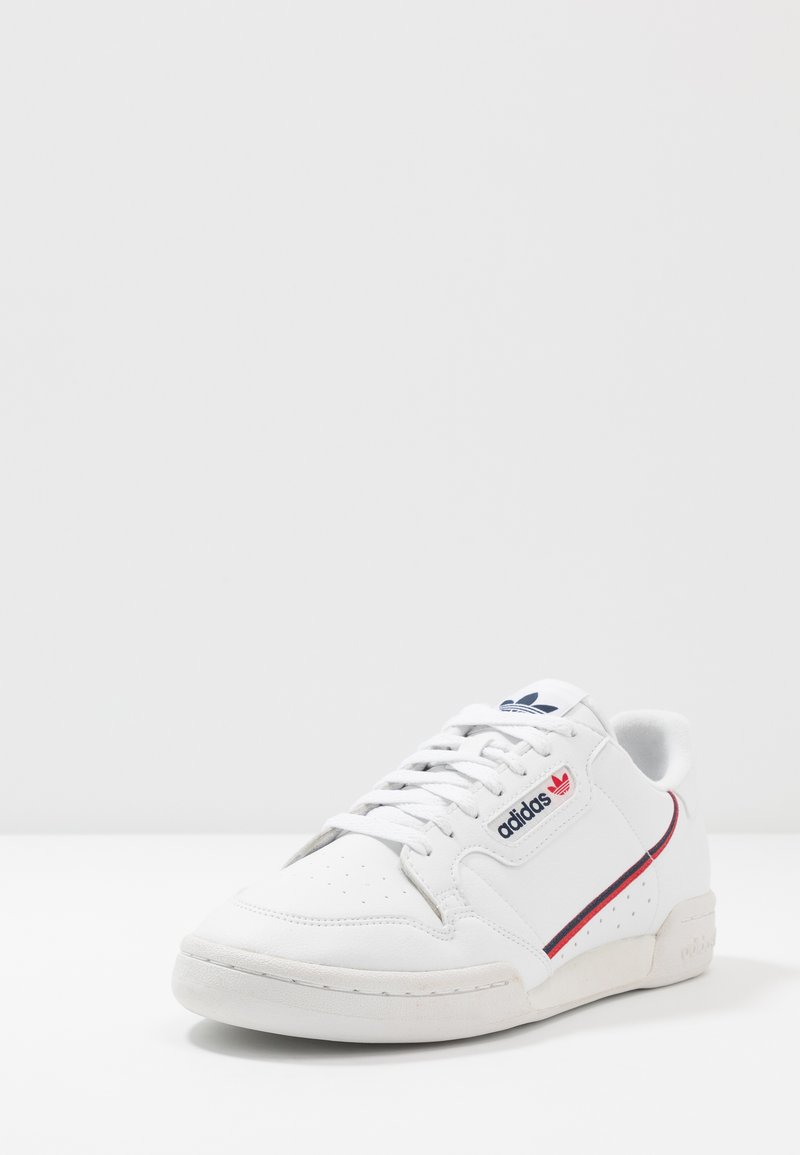 White leather sneaker with perforated toe, flat laces, red and blue side stripe, and signature logo on tongue and side. Rubber sole.