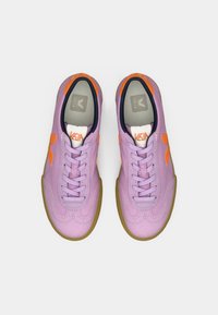 Μωβ αθλητικά παπούτσια με πορτοκαλί λεπτομέρειες, στρογγυλή μύτη και υφή στην επιφάνεια. Διαθέτουν κορδόνια και σόλα από καουτσούκ για καλύτερη πρόσφυση.