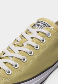 Κίτρινο αθλητικό παπούτσι Converse All Star με λευκά κορδόνια, μεταλλικές τρύπες και ορατή ετικέτα μάρκας στη γλώσσα.