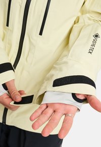 Hellgelbe GORE-TEX-Jacke mit schwarzen Akzenten, ausgestattet mit einem Reißverschluss vorne, Klettverschlussmanschetten und offenen Ärmeln mit Innenfutter, das detaillierte Nähte zeigt.