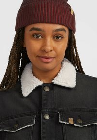 Sort denimjakke med en hvid, blød krave, der har metal snaplukninger og to brystlommer. Båret med en maroon ribbet beanie.