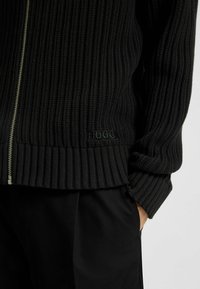 Maglione zip-up a coste nere con logo "HUGO" sull'orlo, indossato con pantaloni neri, mano nella tasca visibile sul lato destro.