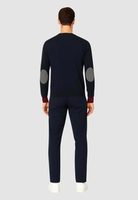 Pullover navy con polsini a costina, presenta toppe grigie sui gomiti, scollatura tonda e design aderente. Abbinato a pantaloni scuri e scarpe bianche.
