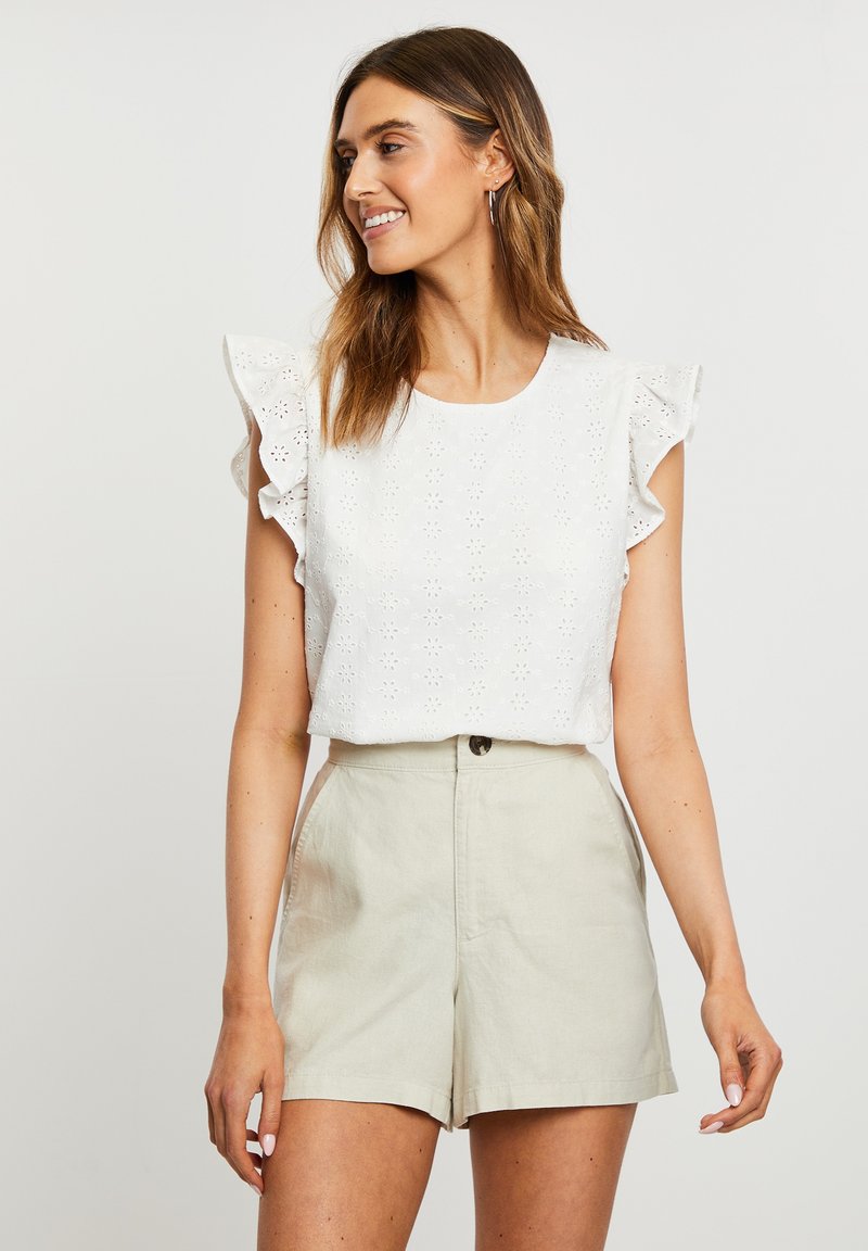 Weiße Bluse mit Lochmuster und Rüschensäumen, kombiniert mit hellbeigen hoch taillierten Shorts. Sie hat Blumenmuster und eine weiche Textur.