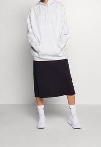 Personne portant un hoodie blanc Calvin Klein, une jupe midi noire, des chaussettes blanches Adidas et des baskets blanches Adidas se tenant sur un sol gris.