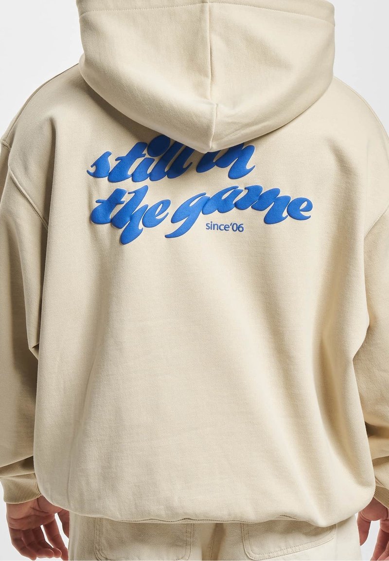 Beige mikina s modrým nápisem "stále v hře od '06" na zadní straně, nošena osobou obrácenou zády.
