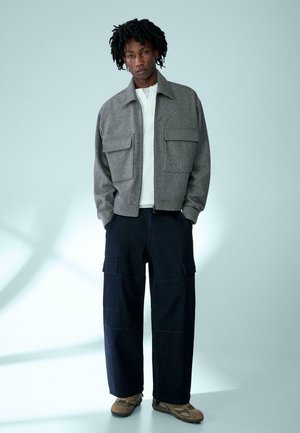 Man standing with hands in pockets wearing a gray zip jacket, white shirt, wide dark cargo pants, and brown sneakers against a plain background.