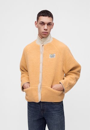 Un mannequin se tient devant un fond uni, vêtu d’une veste sherpa couleur pêche avec des finitions crème et d’un jean bleu foncé.