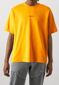 Světlý oranžový bavlněný tričko s kulatým výstřihem a krátkými rukávy. Na hrudi se nachází malé černé logo "G-STAR RAW".