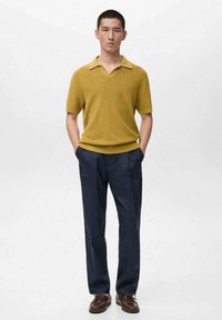 Polo a maniche corte color senape con scollo a V, abbinata a pantaloni sartoriali blu navy scuro e scarpe casual marroni. Design semplice, tessuto strutturato.