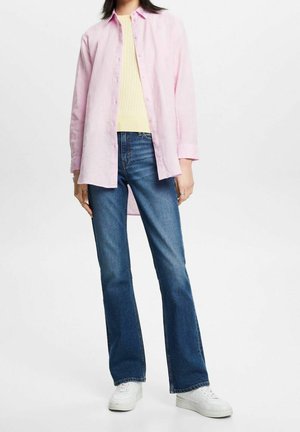 Persona vistiendo una camisa de botones color rosa claro sobre un suéter amarillo pálido, jeans azules y zapatillas blancas, de pie contra un fondo blanco.