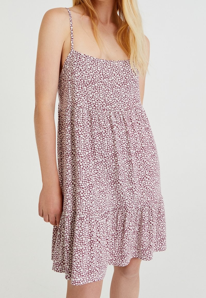 Femme portant une robe d'été sans manches rose et blanche à motifs floraux avec fines bretelles et ourlet volanté, debout devant un fond uni.
