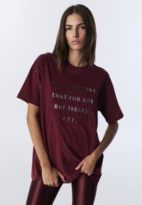 Burgundowa bawełniana koszulka z krótkim rękawem i luźnym krojem, z odblaskowym napisem na środku, który zawiera motywującą frazę.