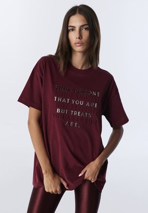 Burgundowa bawełniana koszulka z krótkim rękawem i luźnym krojem, z odblaskowym napisem na środku, który zawiera motywującą frazę.