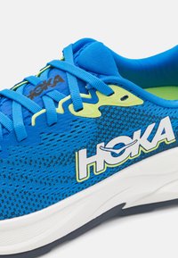 Blauwe hardloopschoen met bovenwerk van mesh, contrasterende limoenaccenten, HOKA-logo en witte tussenzool. Heeft een gestructureerd oppervlak en ronde vorm.