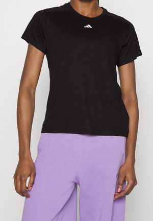 Persoon die een effen zwart T-shirt met korte mouwen draagt met een klein wit logo en lichtpaarse broek, staand tegen een effen achtergrond.