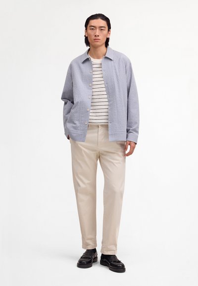 Junger Mann steht und trägt eine blaue karierte Jacke über einem weiß-schwarz gestreiften Hemd, beigefarbene Hosen und schwarze Schuhe vor einem weißen Hintergrund.