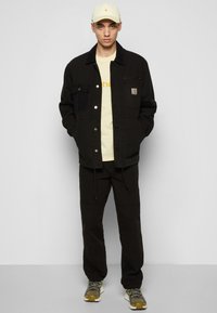 Casaco de corduroy preto com bolsos frontais, t-shirt bege com texto laranja, calças pretas e sapatilhas multicoloridas. Um boné claro completa o visual.