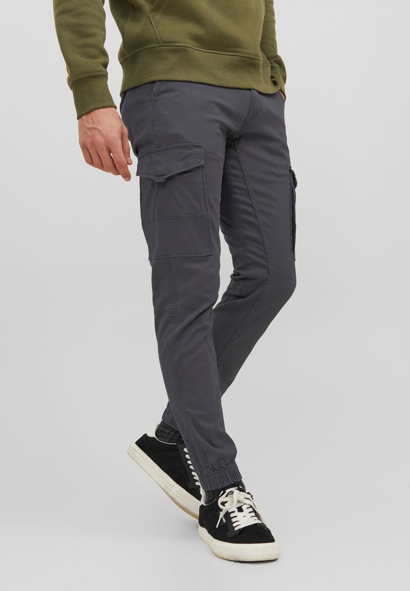 Pantalones cargo grises con puños elásticos, bolsillos laterales y una textura suave. Combinados con zapatillas negras que tienen cordones blancos y una suela texturizada.