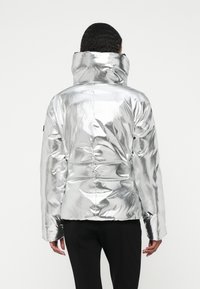 Casaco acolchoado prateado com um colarinho alto, apresentando uma superfície brilhante e refletiva, detalhes franzidos na parte de trás e mangas longas.