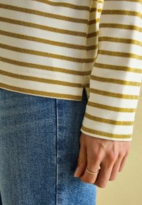 Gestreiftes Langarmoberteil in Creme und Gold mit gerippter Textur; kombiniert mit blauen Denim-Jeans. Die Ärmel zeigen einen seitlichen Schlitz und einen subtilen metallic Faden.