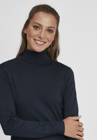 Námornícky rolák z rebrovanej pleteniny, s dlhými rukávmi a priliehavými manžetami. Model sa usmieva na neutrálne pozadí.