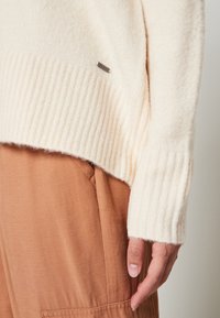 Maglione a maglia di colore crema chiaro con polsini e orlo a coste, caratterizzato da un piccolo etichetta brandizzata in argento. Indossato sopra pantaloni in tessuto color ruggine.