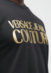 Melnas krāsas krekls ar lieliem zelta metāliskiem burtiem, kas lasāmi "Versace Jeans Couture", valkāts cilvēka.