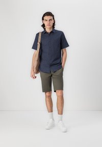 Dunkelblaues kurzärmliges Hemd mit einem dezenten Muster, kombiniert mit olivgrünen Shorts. Weiße Sneakers und eine sandfarbene Umhängetasche runden den Look ab.