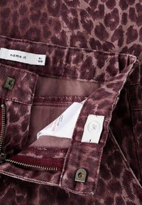 Tuvumā redzama bordo leopardu raksta džinsaudums ar rāvējslēdzēju, pogu un redzamām apģērba etiņām iekšējā jostasvietā.