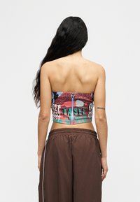 Femme aux longs cheveux noirs portant un crop top graphique sans bretelles avec une fermeture éclair et un pantalon marron ample, vue de dos.